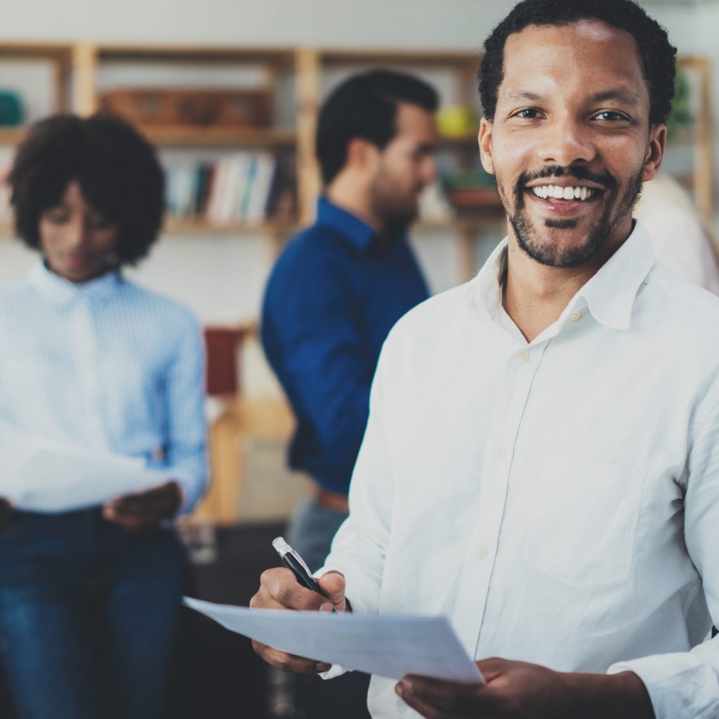 Young african american businessman wearing white shirt holding papers at hands and standing front of the coworkers team.Horizontal,blurred background.