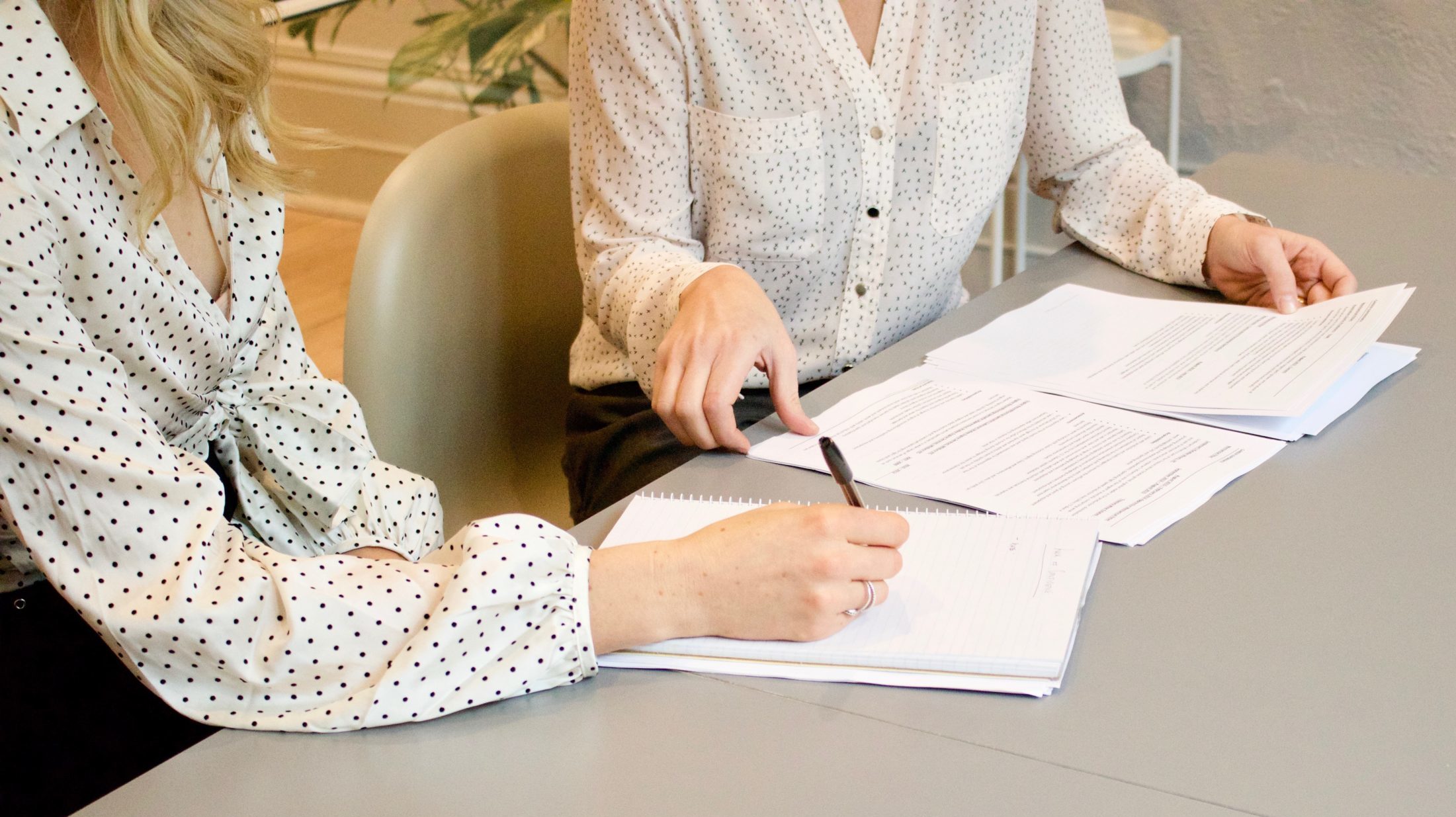 woman signing on white printer paper beside woman about to touch the documents